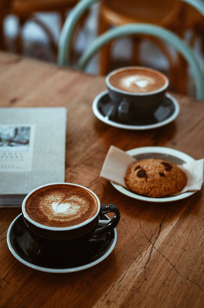 Cozy cafe setup with cappuccinos and a chocolate chip cookie on a rustic wooden table.