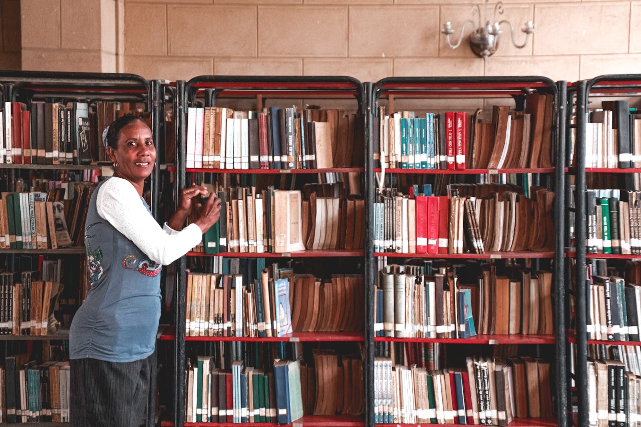 Side view of content mature Hindu female picking textbook from bookcase with assorted literature while looking at camera in library