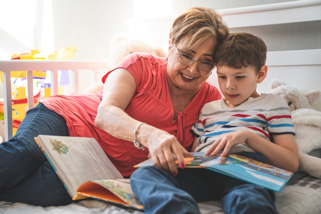 Elderly woman reading a book with her grandson on a cozy bed in a sunlit room.